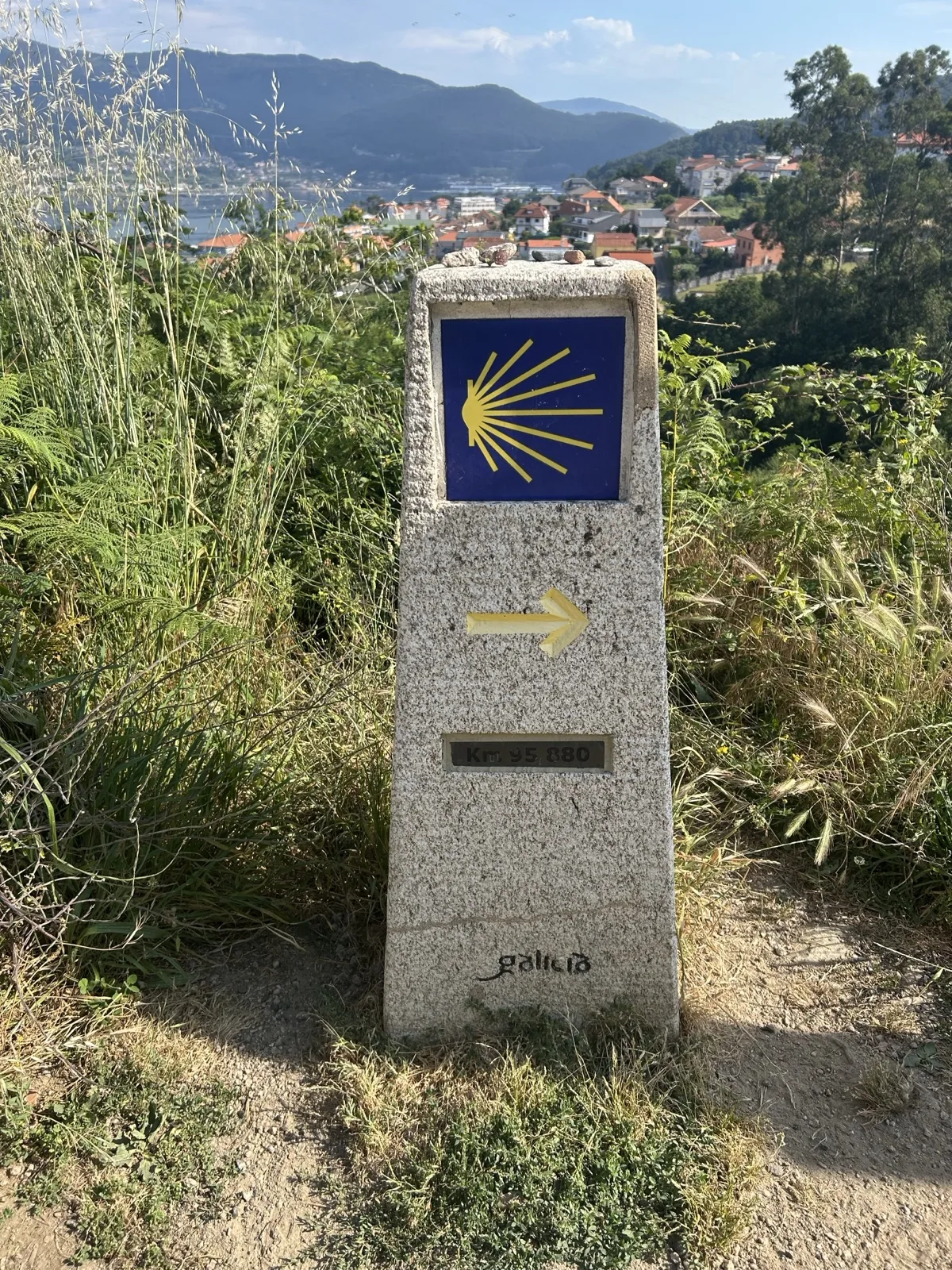 Wegstein des Jakobswegs in Galicien mit Muschel und gelbem Pfeil, dahinter eine Bucht mit Häusern
